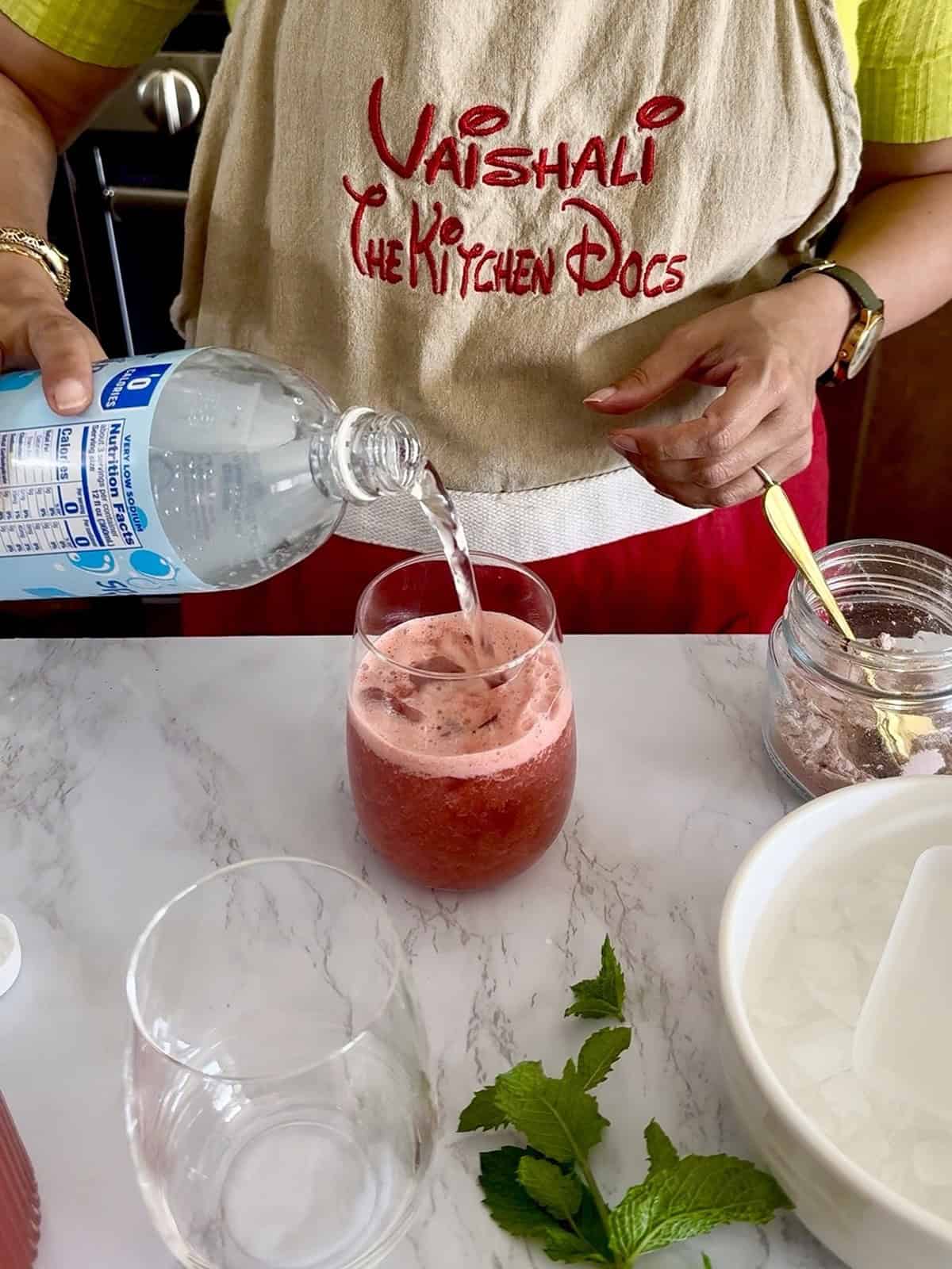 Pouring fizzy sparkling water over a chilled pink watermelon juice base with ice cubes to create a summer mocktail.