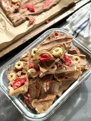 Nut-free high protein chocolate yogurt bark broken into pieces in a freezer safe glass container with SunButter swirl and crispy cereal and strawberry topping.