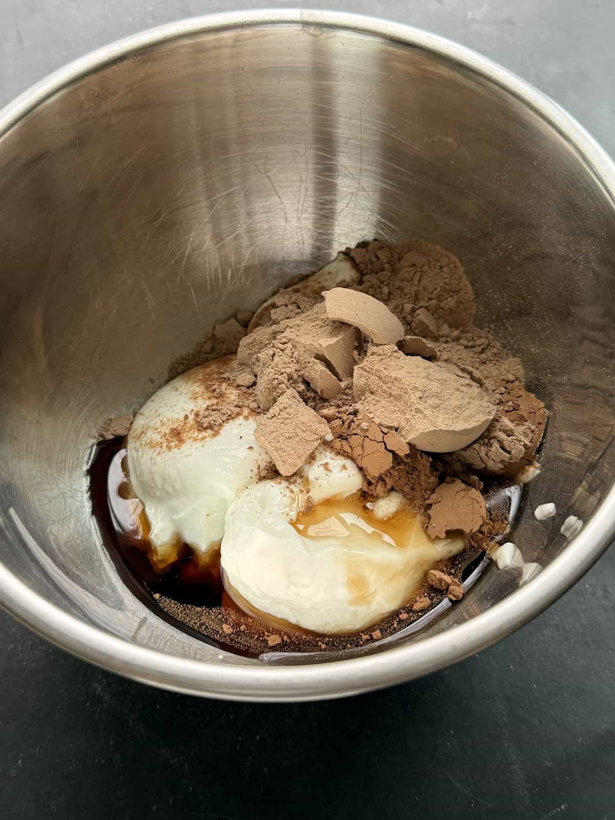 Maple syrup, greek yogurt, chocolate protein powder, and cocoa powder in a stainless steel mixing bowl.