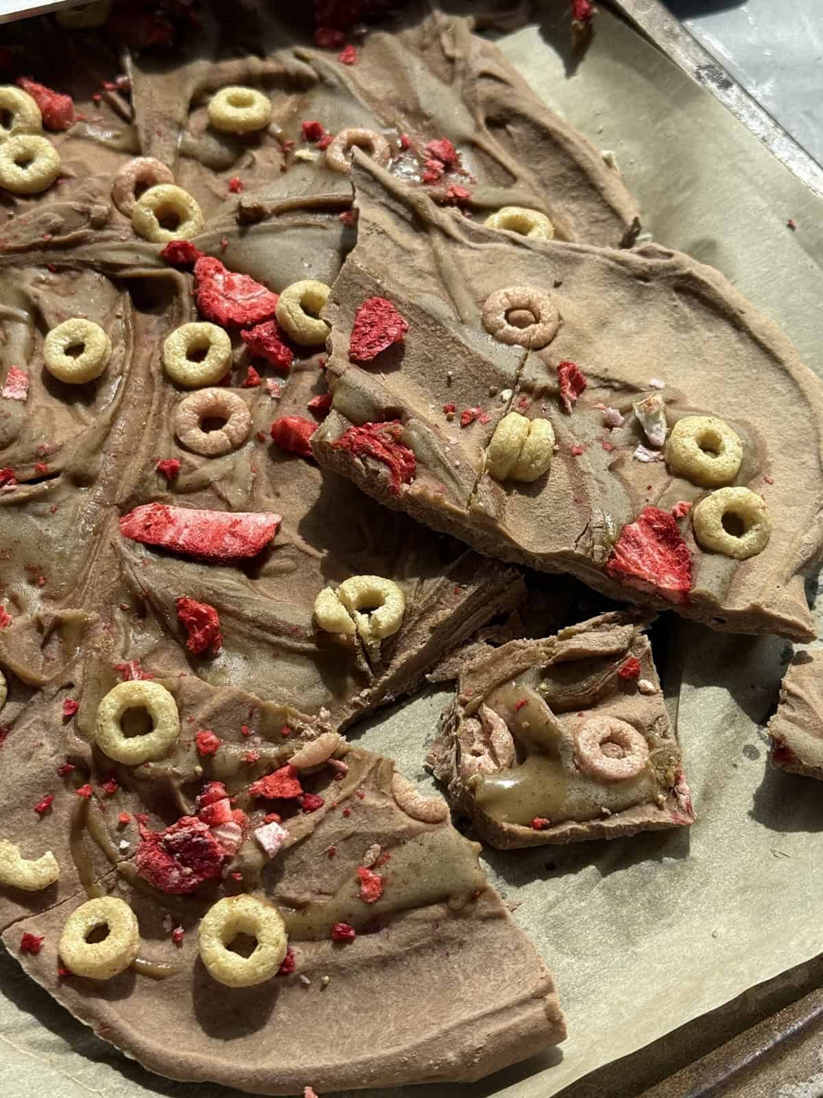 Nut-free high protein chocolate bark broken into pieces on parchment paper with SunButter swirl and crispy cereal and dried strawberry topping.