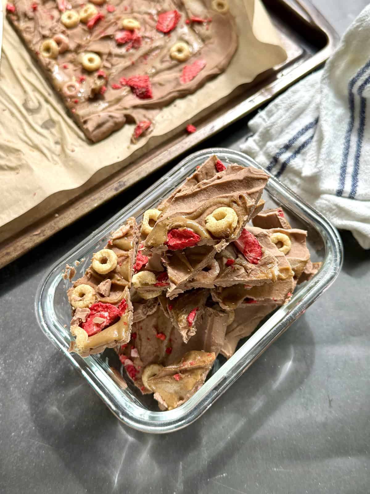 Nut-free high protein chocolate yogurt bark broken into pieces in a freezer safe glass container with SunButter swirl and crispy cereal and strawberry topping.