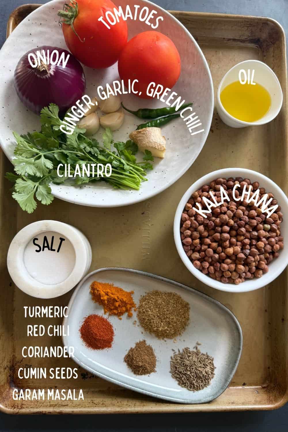 Labeled ingredients for kala chana curry including dry black chickpeas, onion, tomato, ginger, garlic, green chili, and Indian spices laid out on a kitchen tray