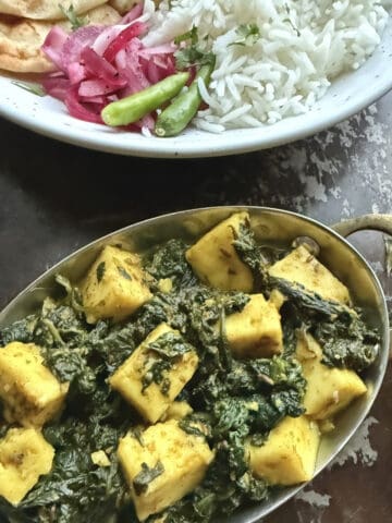 Easy palak paneer without a blender served in a traditional Indian dish. A white ceramic dish in the background has cooked rice, naan, and pickled onions to serve this palak paneer with.