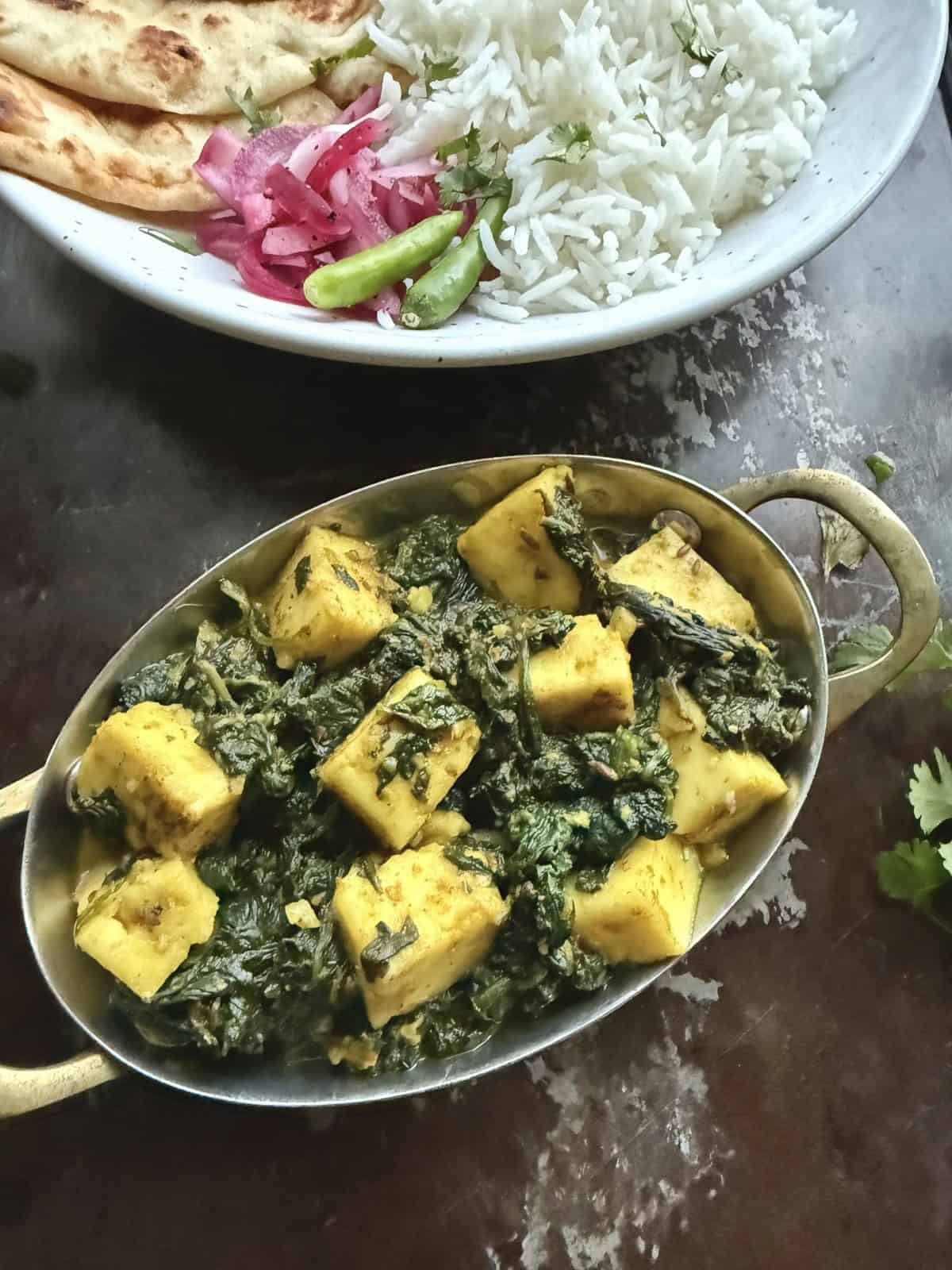 Delicious palak paneer curry served in a traditional Indian dish. A white ceramic dish in the background has cooked rice, naan, and pickled onions to serve this palak paneer with.