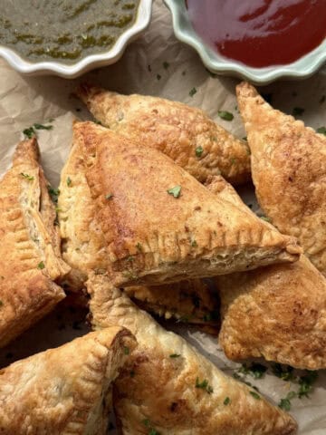 Aloo puff recipe - crispy golden Indian bakery style potato puff pastry on a parchment lined board with green and red chutneys.